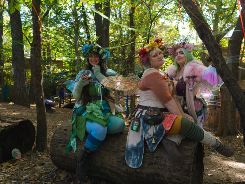Three women dressed as colorful fairies sit on a log.