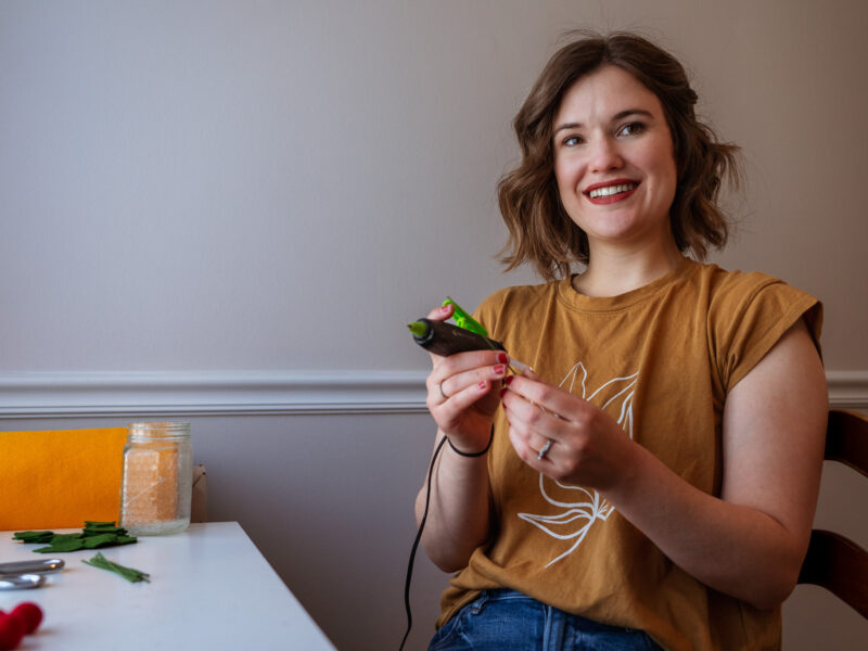 A woman holds a hot glue gun and smiles at the photographer