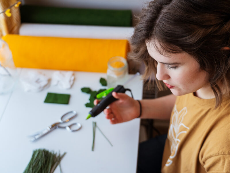 A woman holds a glue gun and looks down at her desk.