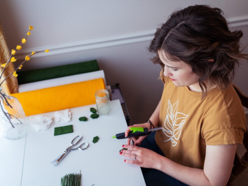 A woman hot glues green felt at her desk.