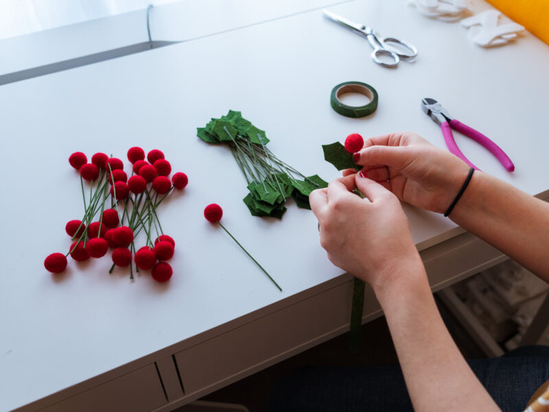 Hands make felt holly bunches at a desk.