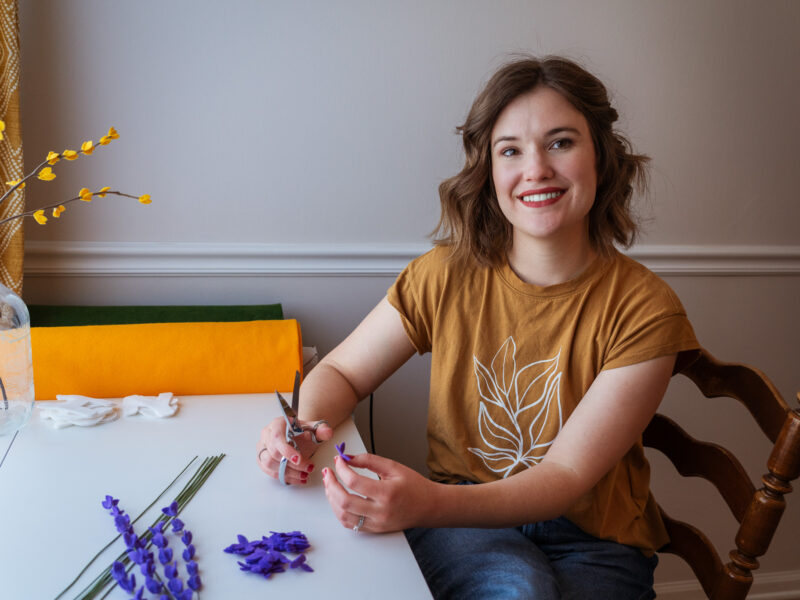 A woman smiles at the camera while making lavender felt stems.